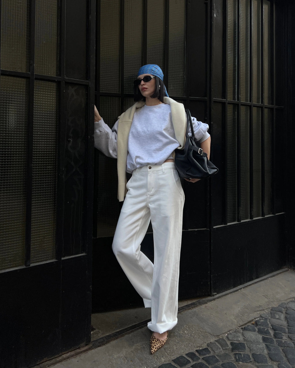 Person wearing a white outfit with a blue hat and carrying a black bag against a dark wall.Wearing beige jeans.