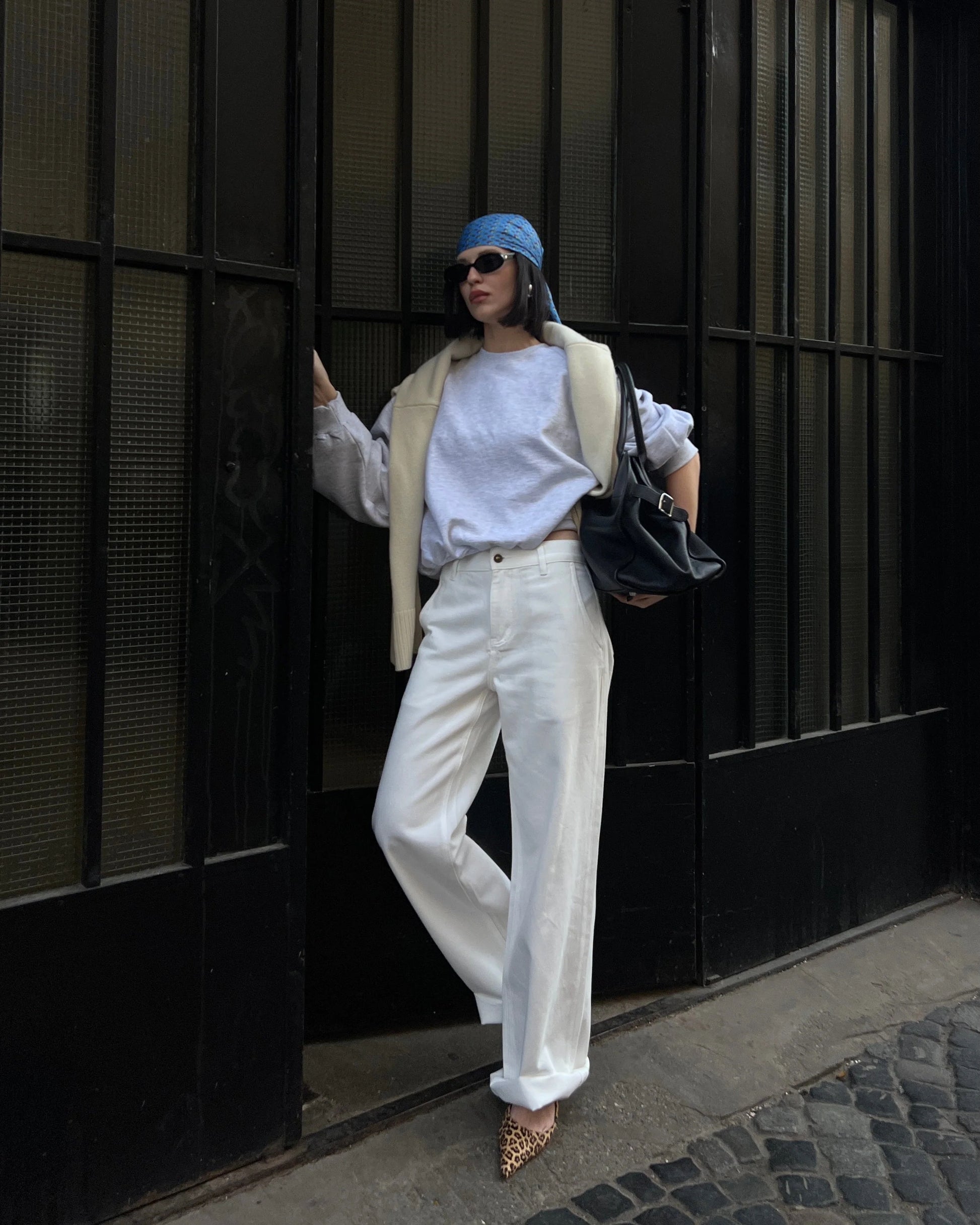 Person wearing a white outfit with a blue hat and carrying a black bag against a dark wall.Wearing beige jeans.